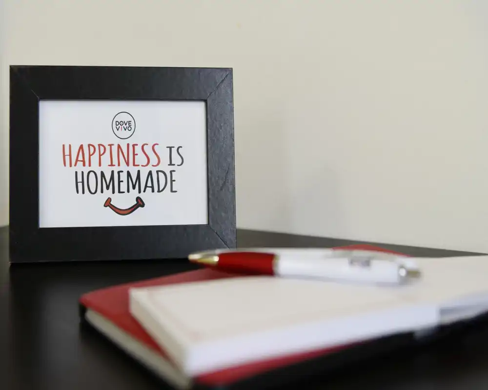 Close-up of a tabletop vignette with a framed sign reading "Happiness is homemade", a pen and an open notebook. The composition suggests a decorative desk or shelf detail rather than a full room view.