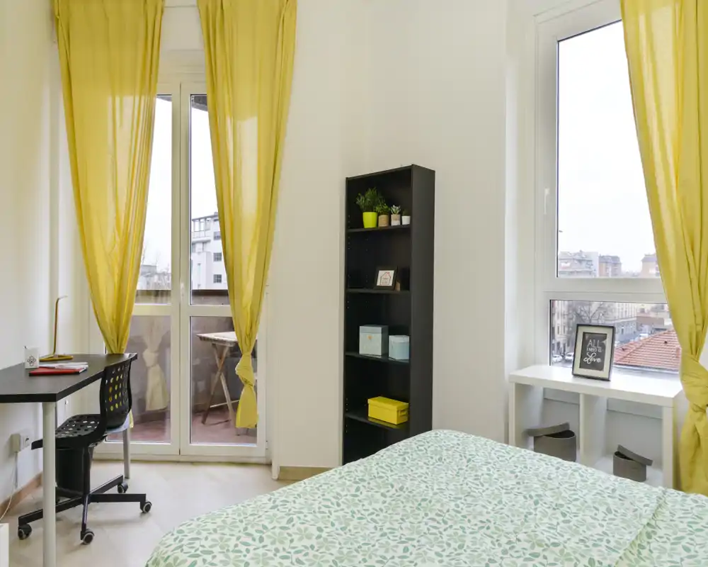 Bright double bedroom with large windows, yellow curtains and a patterned bedspread; includes a desk by the balcony door and shelving for storage — airy and welcoming.