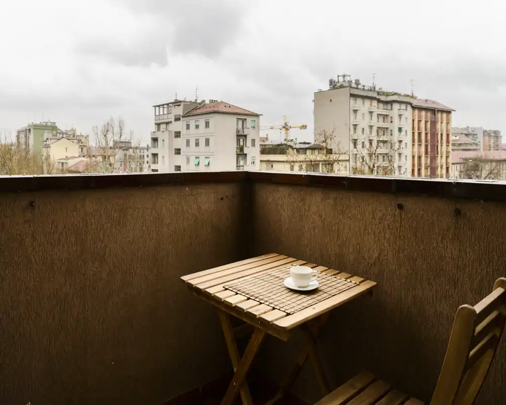 Cozy urban balcony with a small wooden table and chair, ideal for coffee while overlooking neighboring buildings on an overcast day.