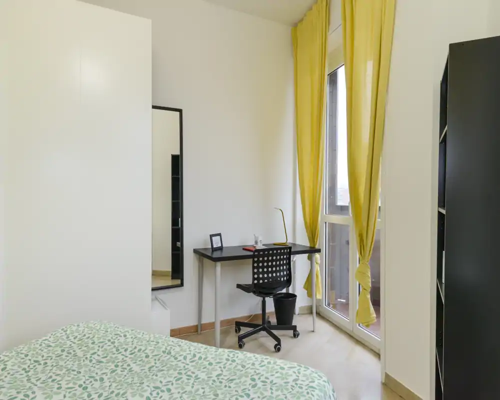 Bright, modern bedroom with patterned bedspread, a compact desk by the window and tall yellow curtains that add color and natural light.