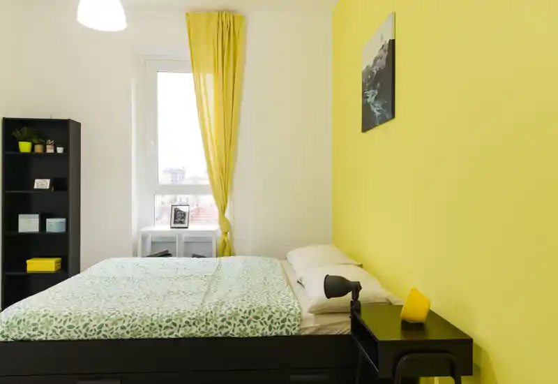 Sunny, modern bedroom with a double bed, patterned green bedding and a bright yellow accent wall. The room features a window with yellow curtains, a bedside table and shelving for storage.