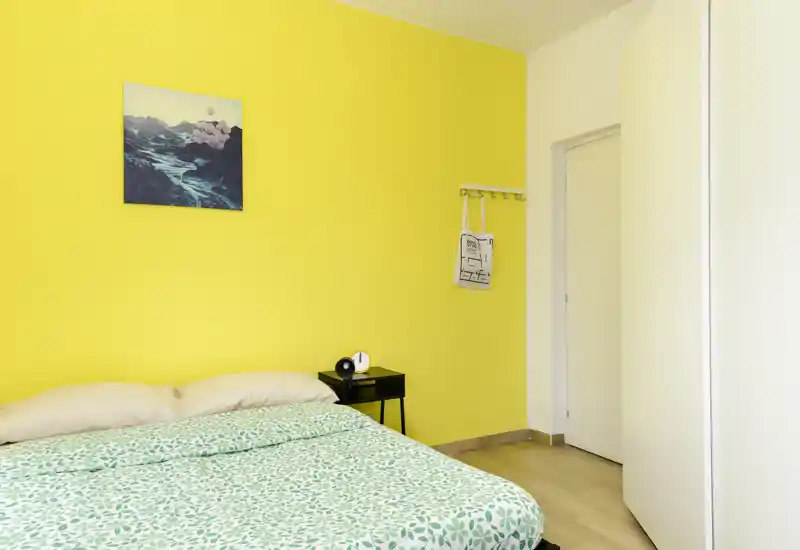 Bright, minimal bedroom with a green patterned duvet against a bold yellow wall, bedside table and simple décor — airy and modern.