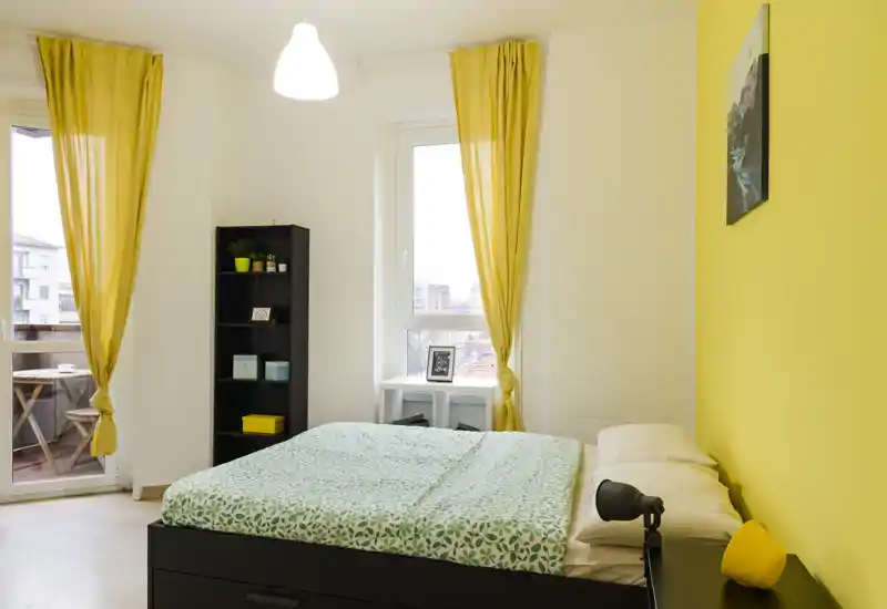 Bright, modern bedroom with a double bed, yellow accent wall and matching curtains, plus a small balcony visible through a glass door.