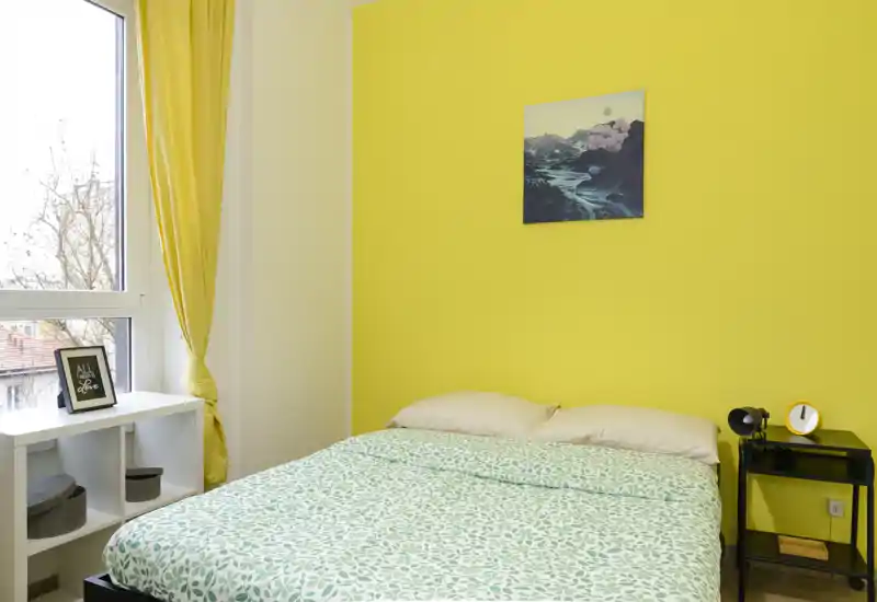 Sunny bedroom with a double bed against a vibrant yellow wall, patterned duvet, bedside table with lamp and clock, and a bright window that fills the room with natural light.