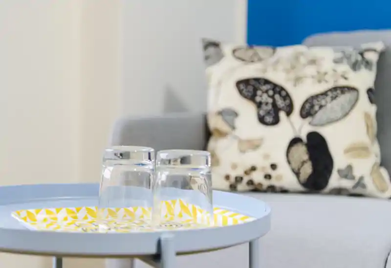 Primer plano de una mesa auxiliar del salón con dos vasos sobre una bandeja decorativa y un cojín estampado en el sofá cercano, todo con luz suave y natural.