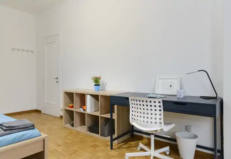 Dormitorio luminoso y minimalista con la estructura de la cama visible, escritorio y estantería. Suelo de parqué, zona de trabajo ordenada con lámpara y pequeños detalles decorativos como una planta.