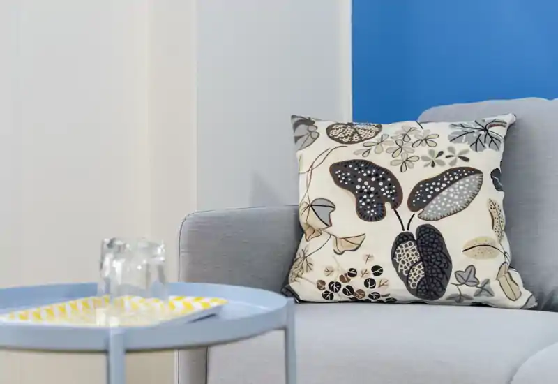 Rincón de salón luminoso y moderno con sofá gris claro, cojín decorativo estampado y una pequeña mesa auxiliar redonda — acogedor y bien decorado para un anuncio.