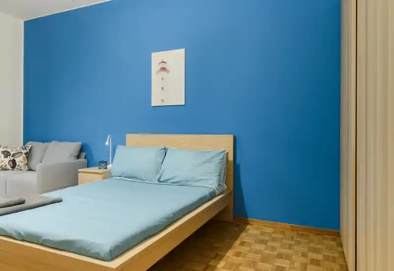Dormitorio moderno y luminoso con cama baja de madera frente a una pared azul intensa, ropa de cama a juego y un pequeño sofá lateral.