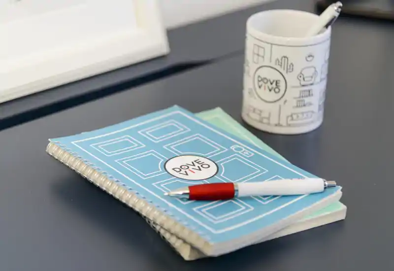 Primer plano de un cuaderno, un bolígrafo y una taza con marca sobre una mesa oscura: foto detalle de papelería y accesorios de escritorio.