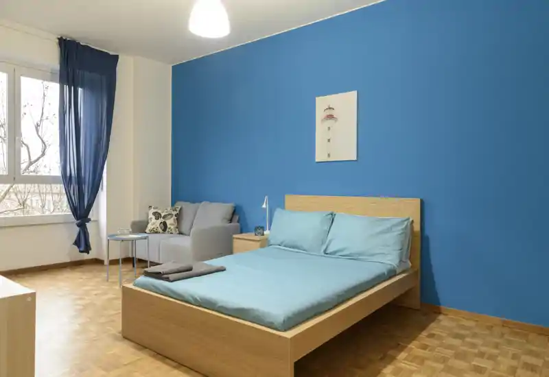 Camera da letto moderna e luminosa con letto in legno su base piatta davanti a una parete blu intensa, biancheria turchese e un piccolo angolo salotto vicino alla finestra.