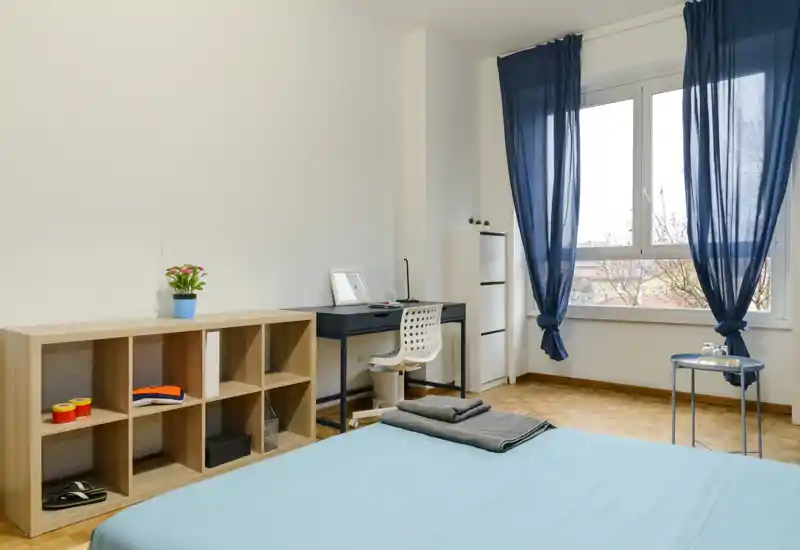 Camera da letto luminosa e minimal con letto ben rifatto, scrivania per lo studio e ampia finestra con tende blu che lasciano entrare luce naturale.
