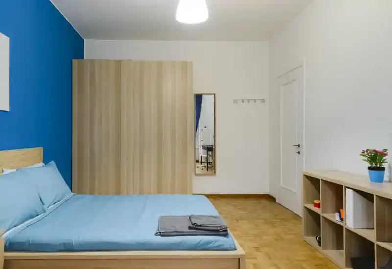 Camera da letto luminosa e moderna con letto matrimoniale in lenzuola blu, armadio in legno, scaffalatura e specchio a parete — stile pulito e minimal, ideale per annunci.