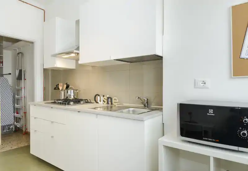 Cocina compacta y moderna con muebles blancos, placa de gas, fregadero integrado y microondas en una estantería lateral — luminosa, limpia y práctica para alquileres de corta estancia.