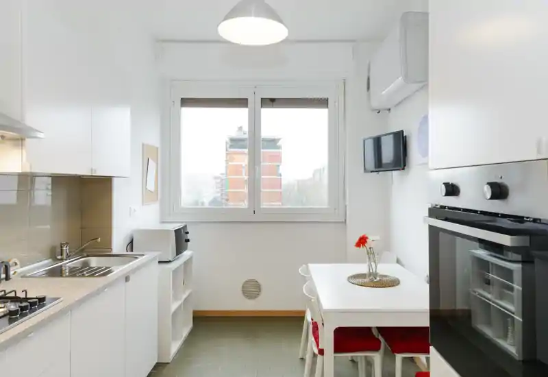 Cocina compacta y luminosa con muebles blancos, horno integrado y placa de gas, pequeña mesa con cojines rojos y ventana con vista a la ciudad — ideal para un apartamento compacto.