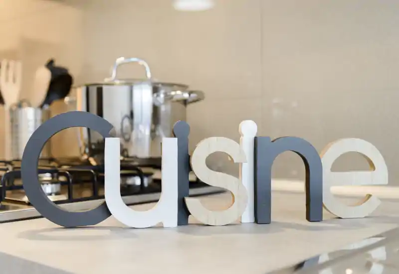 Primer plano de una encimera de cocina moderna con cocina y letras decorativas de madera que leen “Cuisine”, mostrando una zona de cocinado luminosa y con estilo.