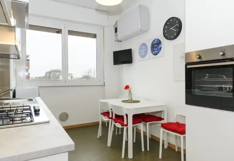 Cocina compacta y luminosa con ventana, horno empotrado, placa de gas y una pequeña mesa con cojines rojos — ideal para comidas acogedoras.