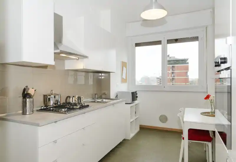 Cocina moderna y luminosa con muebles blancos, placa de gas, fregadero y un pequeño comedor junto a la ventana con una flor decorativa.