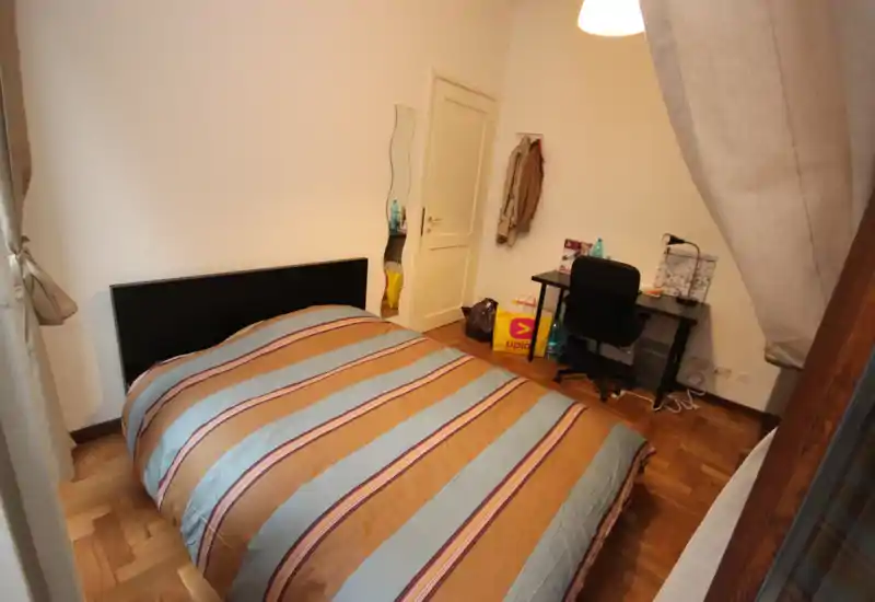Camera da letto accogliente con letto matrimoniale, copriletto a righe, pavimento in parquet e un piccolo angolo studio con scrivania e sedia. Luce calda e pareti neutre rendono l’ambiente confortevole.