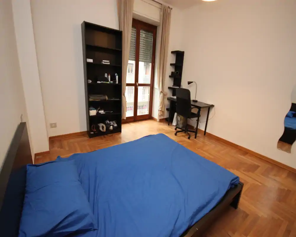 Camera da letto luminosa e ordinata con letto matrimoniale con lenzuola blu, scaffale e una piccola scrivania vicino alla finestra che offre luce naturale.