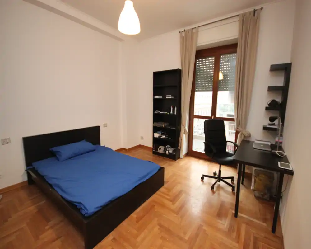 Camera da letto luminosa e essenziale con letto matrimoniale, pavimento in parquet, scrivania e scaffalatura vicino a una grande finestra/porta che lascia entrare luce naturale.