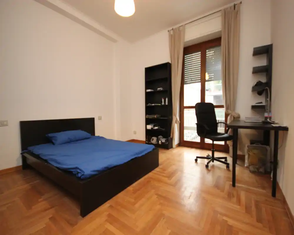 Camera da letto semplice e luminosa con letto matrimoniale, pavimento in parquet e scrivania accanto alla porta finestra. Ampia luce naturale e arredi minimal, ideale per uno studente o un professionista.