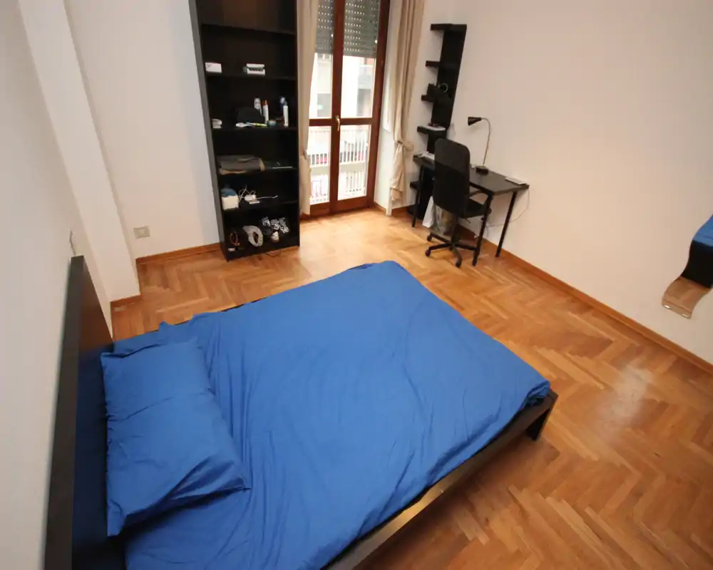 Ampia camera da letto con letto matrimoniale con biancheria blu, parquet e scrivania vicino alla finestra che garantisce luce naturale — semplice, ordinata e pronta all'uso.