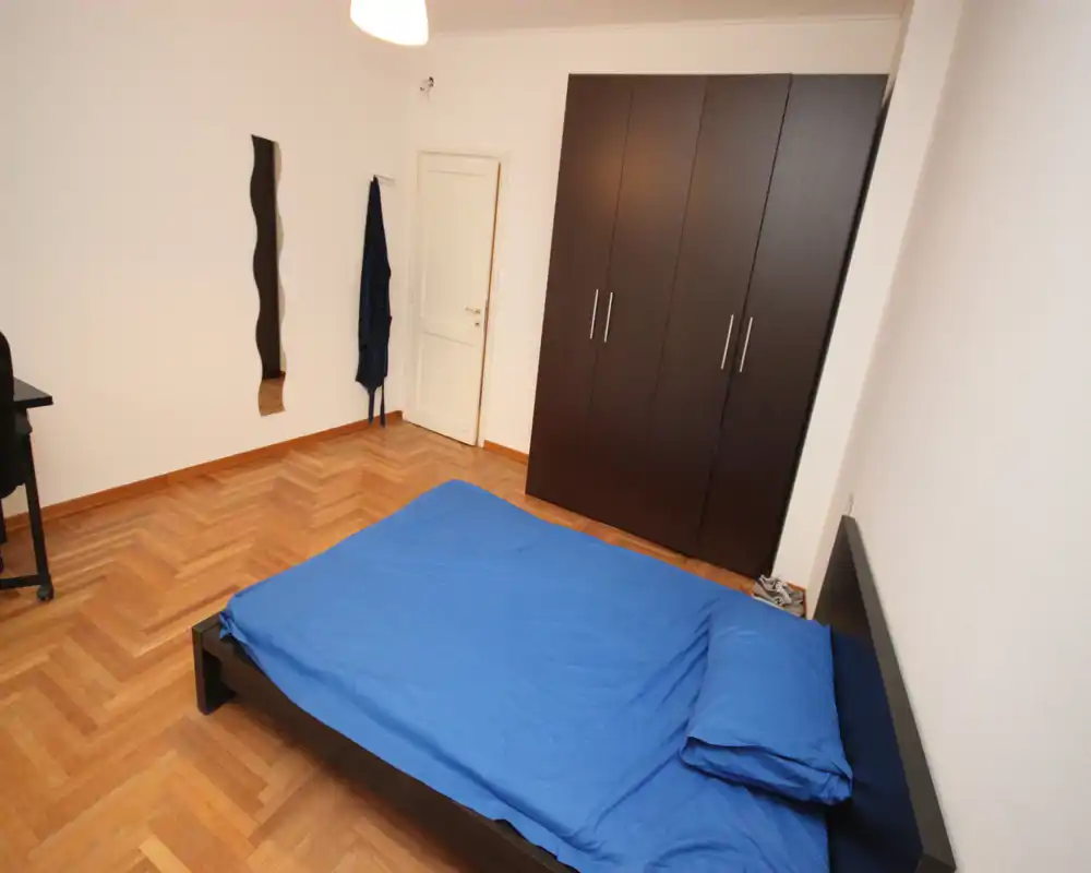 Camera da letto semplice e luminosa con letto matrimoniale con biancheria blu, un grande armadio scuro e pavimento in parquet: arredo minimale e funzionale, adatto per affitti brevi.