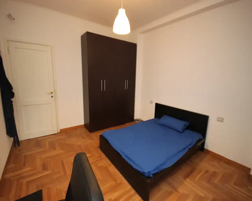 Camera da letto semplice e ben illuminata con letto matrimoniale con biancheria blu, armadio scuro e pavimento in parquet. Layout minimal e ordinato, adatto per affitti brevi.