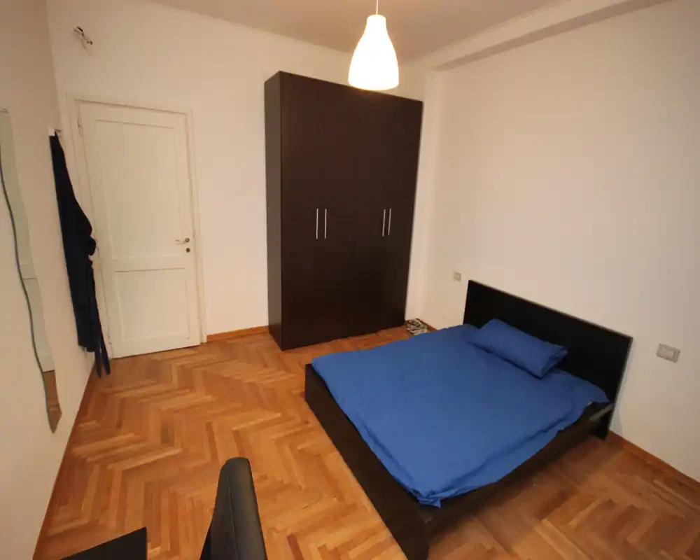 Camera da letto semplice e luminosa con letto matrimoniale contro il muro, un grande armadio scuro e pavimento in parquet; arredo pulito e minimale, ideale per un annuncio.
