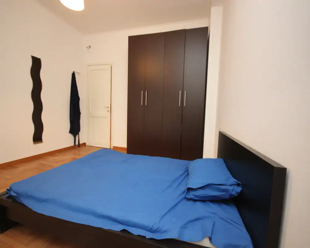 Camera da letto luminosa e moderna con letto matrimoniale con biancheria blu e un ampio armadio scuro. Linee pulite e arredo essenziale creano uno spazio ordinato e tranquillo, ideale per affitti brevi.