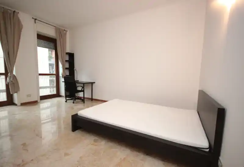 Spacious bedroom with a low dark wooden bed frame and large windows that flood the room with natural light. Simple, modern layout with a small desk and neutral curtains — bright and minimally furnished.