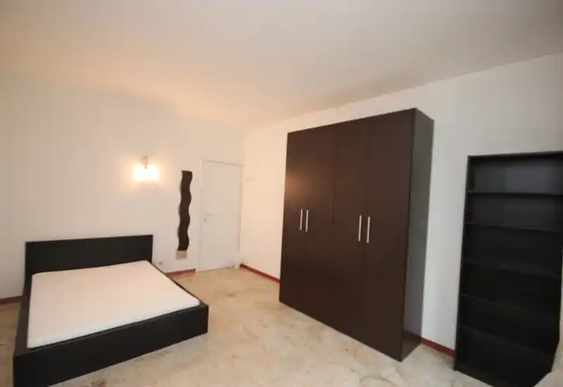 Spacious, simply furnished bedroom with a double bed frame and mattress, a large dark wardrobe and bookshelf. Neutral walls and marble-look floor create a clean, minimalist feel with warm wall lighting.