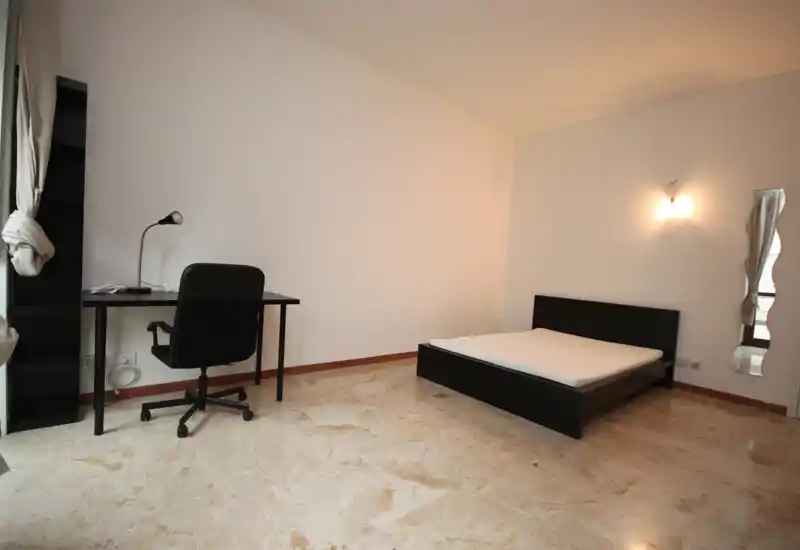 Minimalist bedroom with a double bed on a low dark frame, a desk and office chair by the window, marble floor and soft wall lighting — bright, uncluttered and functional.