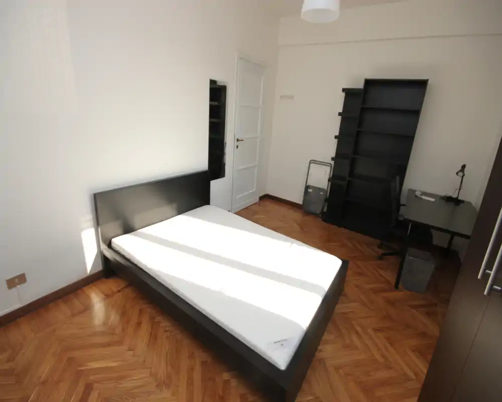 Camera da letto luminosa e minimal con letto matrimoniale su struttura scura, pavimento in parquet e scrivania con scaffale — ideale per uno studente o una persona sola.