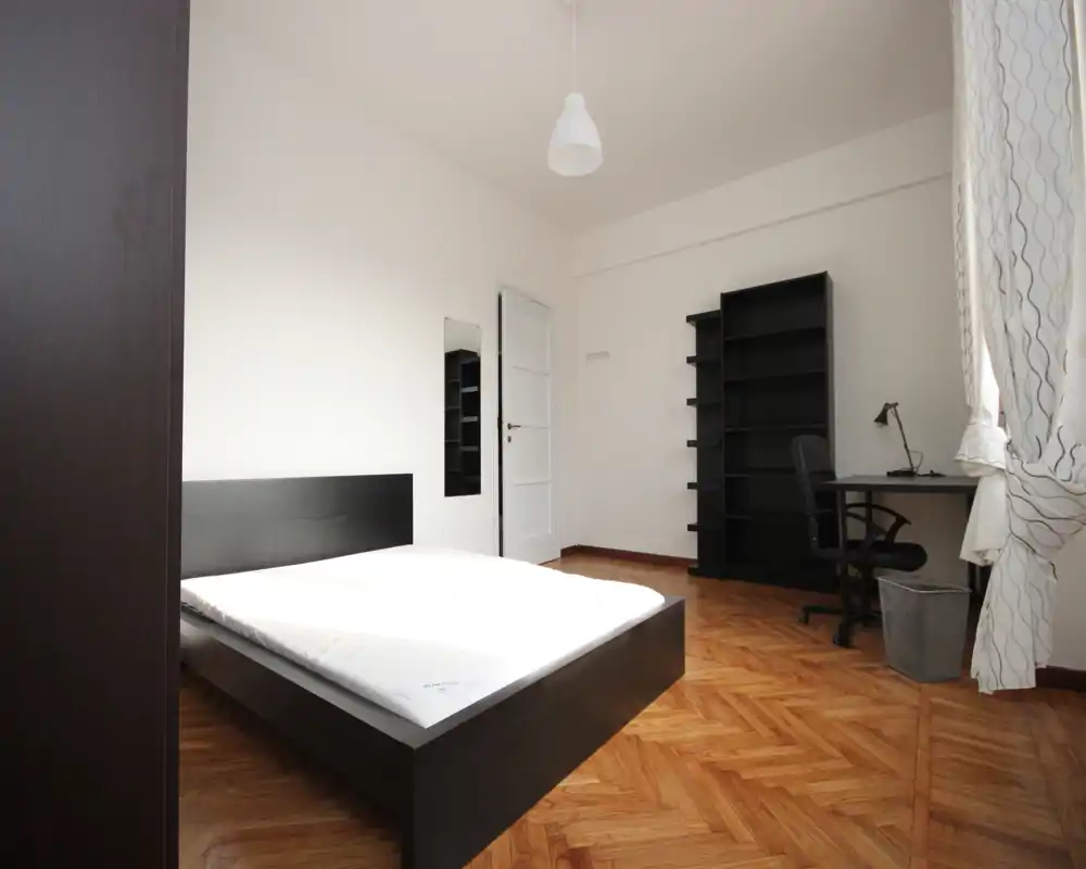 Camera da letto luminosa e moderna con letto basso matrimoniale, pavimento in parquet a spina di pesce, scrivania e scaffalatura vicino alla finestra — ideale per studenti o professionisti.
