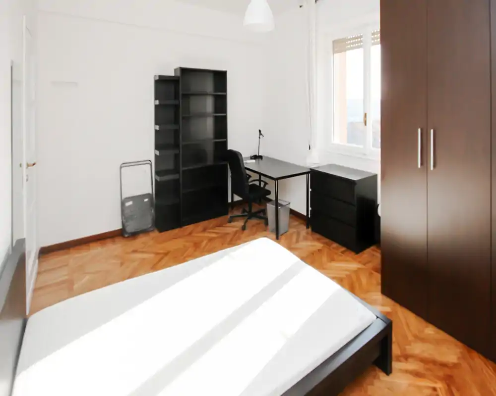 Camera da letto luminosa e ben arredata con letto matrimoniale, pavimento in parquet, armadio e scrivania vicino alla finestra — adatta sia al riposo che al lavoro.