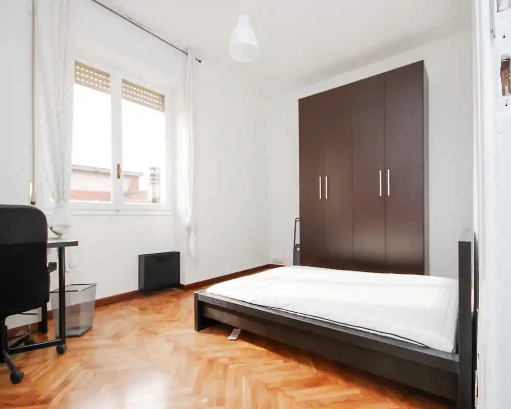 Camera da letto luminosa e minimal con letto basso, grande armadio e pavimento in parquet. La luce naturale dalla finestra crea un ambiente pulito e arioso, perfetto per gli annunci.