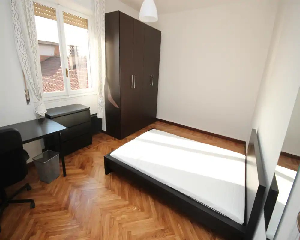 Camera da letto luminosa e minimal con letto basso, armadio e scrivania. Grande finestra con molta luce naturale e pavimento in parquet a spina di pesce caldo.