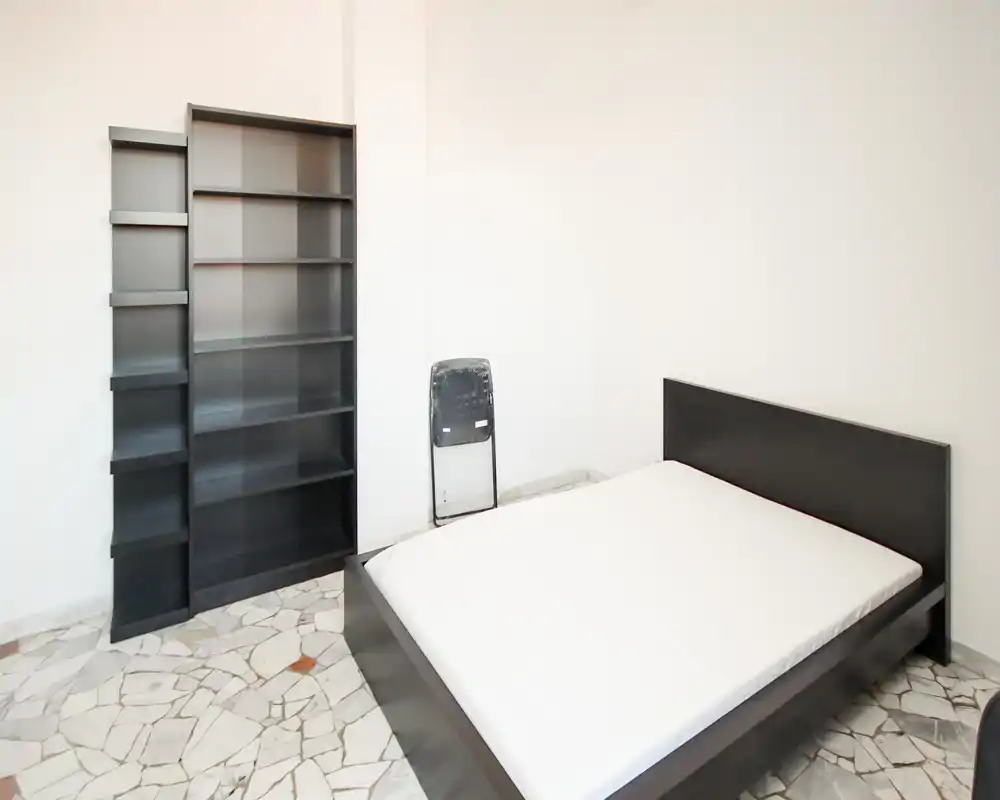 Bright, minimalist bedroom with a double bed on a low dark frame, a tall black shelving unit and tiled floor — tidy, simply furnished and well lit.