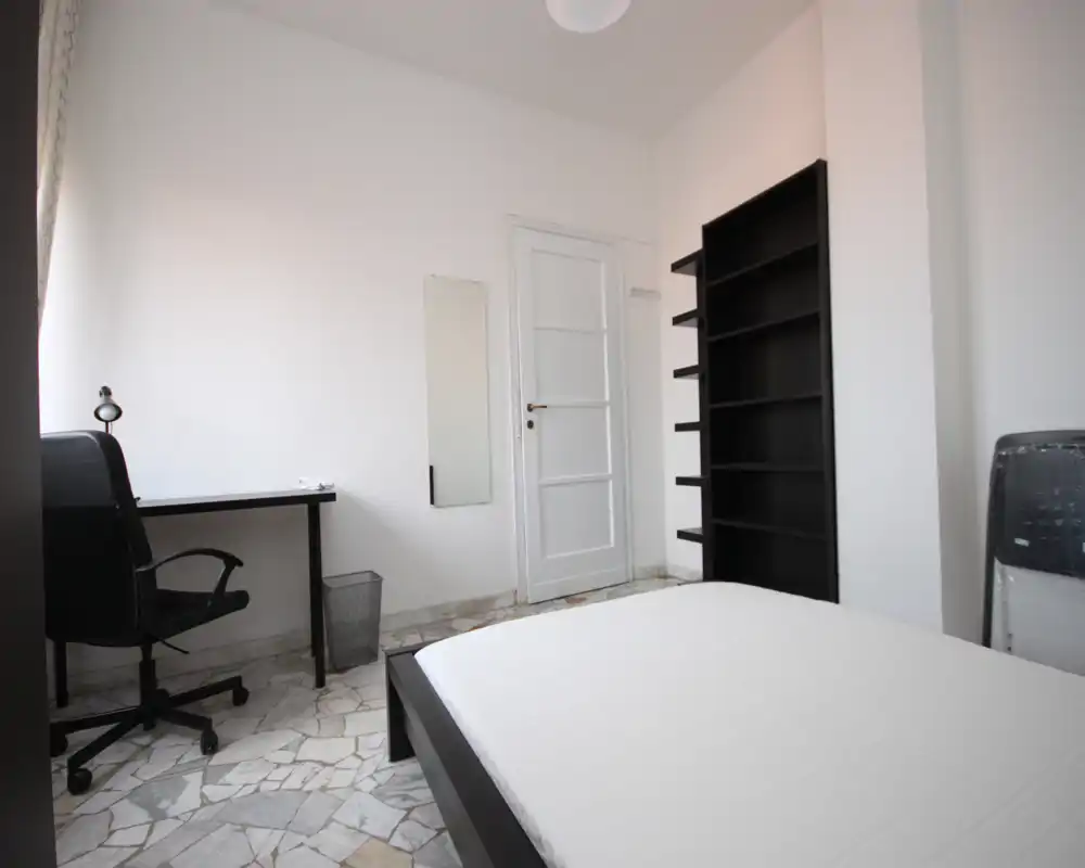 Minimal, well-lit bedroom with a double bed, a desk and chair by the window, and a tall shelving unit. Neutral white walls and marble-patterned floor create a bright, modern feel.