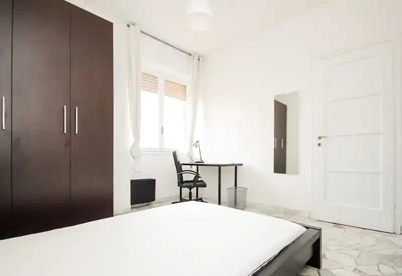 Bright minimalist bedroom with a low platform bed, large dark wardrobe and a small desk by the window — crisp, airy and well-lit space ideal for single occupancy.