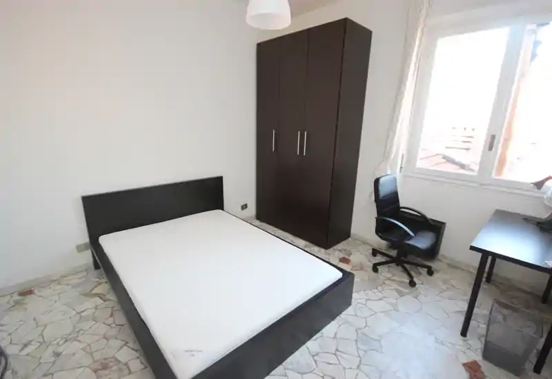 Bright, simply furnished bedroom with a double bed, wardrobe, desk and window providing strong natural light.