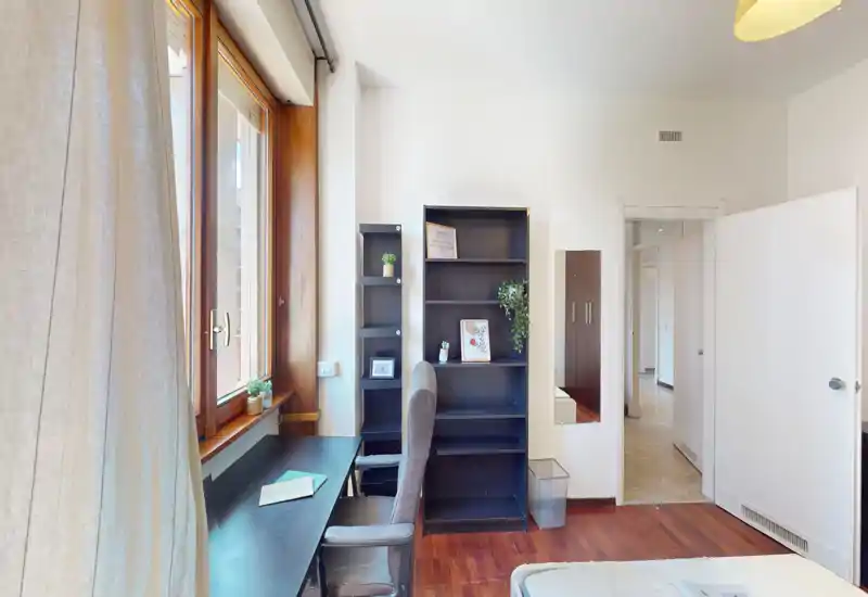 Camera da letto luminosa con angolo lavoro vicino a una finestra con cornice in legno. Si vedono scrivania e sedia, una libreria e parte del letto in primo piano, per un ambiente funzionale e arioso.