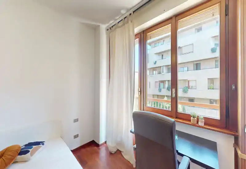 Camera da letto piccola e luminosa con letto singolo, pavimento in legno e ampia finestra; scrivania e sedia vicino alla finestra per luce naturale e vista gradevole.