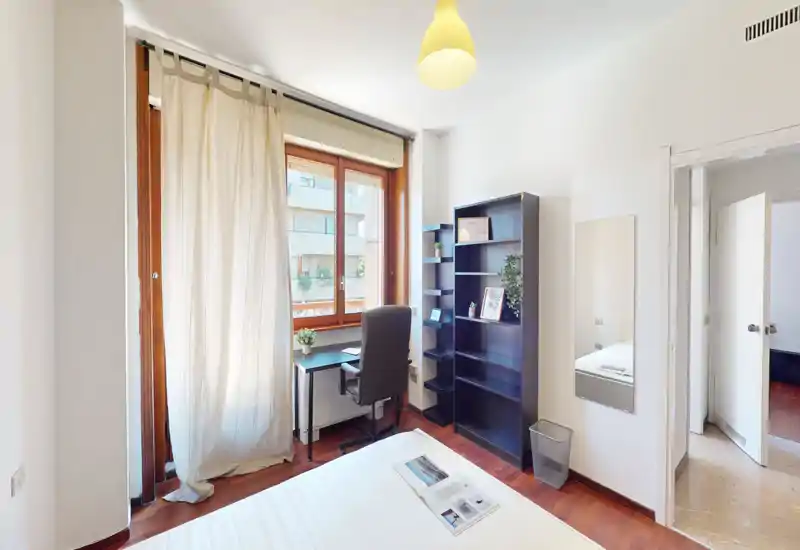 Camera da letto luminosa e moderna con letto comodo, scrivania vicino alla finestra e libreria alta per lo storage — ideale anche per lavorare da casa.