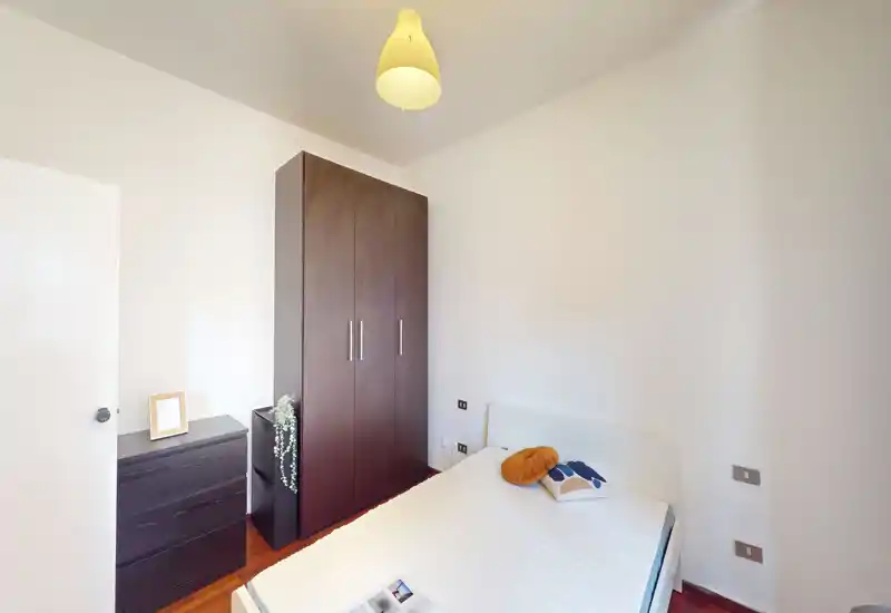 Camera singola luminosa e moderna con letto bianco, cuscini decorativi e un armadio alto in legno scuro; stile semplice e pulito perfetto per un annuncio.