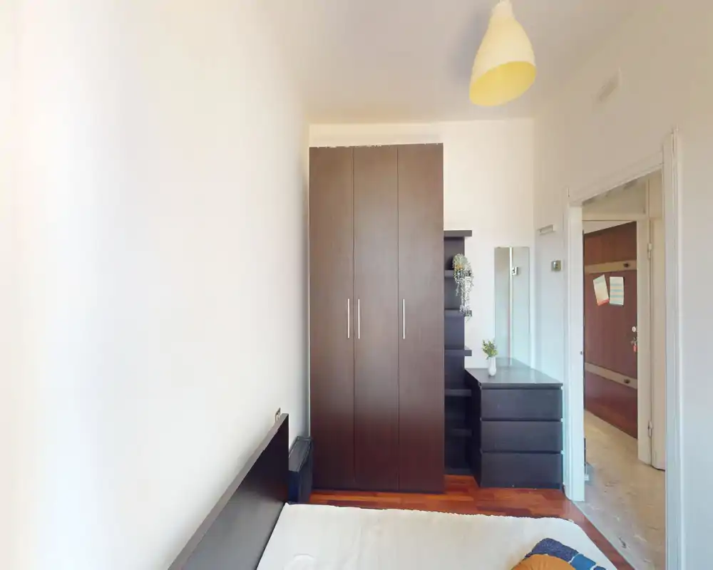 Camera da letto luminosa e compatta con testata del letto e armadio ben visibili. L'arredo è in legno scuro con comodini e cassettiere, ambiente pulito e arioso.