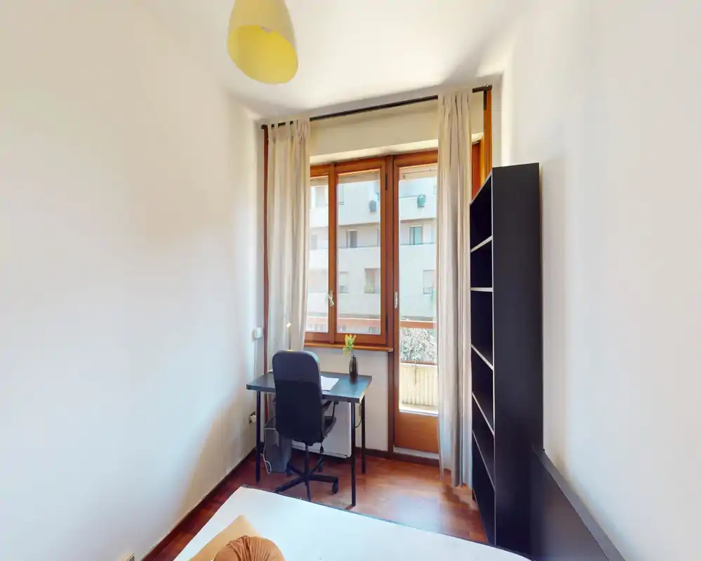 Camera da letto luminosa e minimale con scrivania vicino alla finestra, vista parziale del letto in primo piano e una libreria alta — ideale per una persona o uno studente.