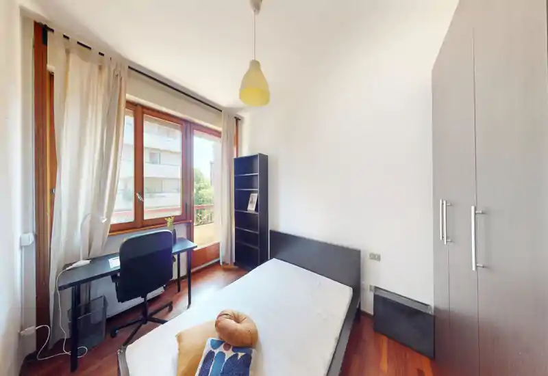 Camera luminosa e arredata in modo semplice con letto singolo, armadio e scrivania vicino alla grande finestra che illumina la stanza.
