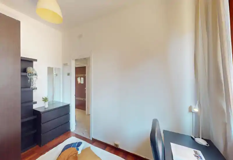 Bright, tidy bedroom corner with a visible bed, dresser and a desk by the window — a practical single-room setup with simple modern furniture and soft natural light.